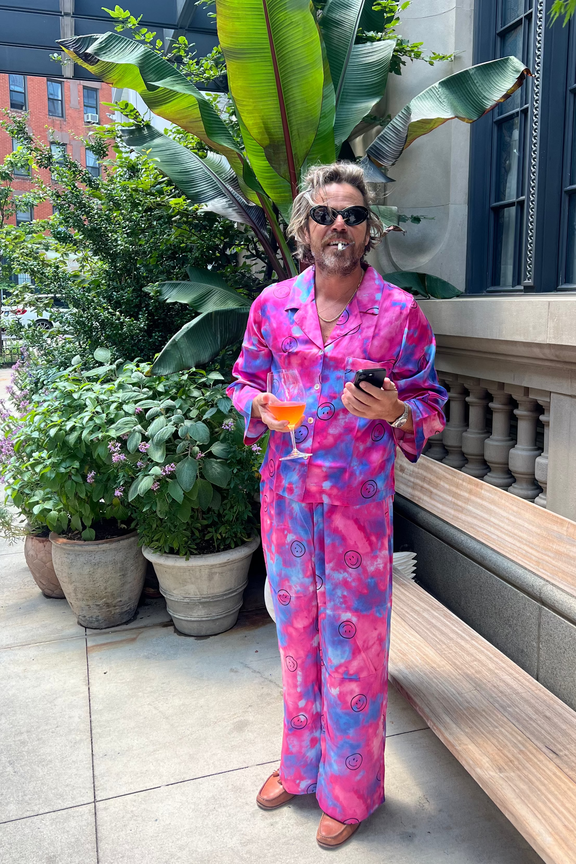 A man wearing a vibrant pink tie dye patterned pajama set stands on an outdoor patio holding a drink and a phone.