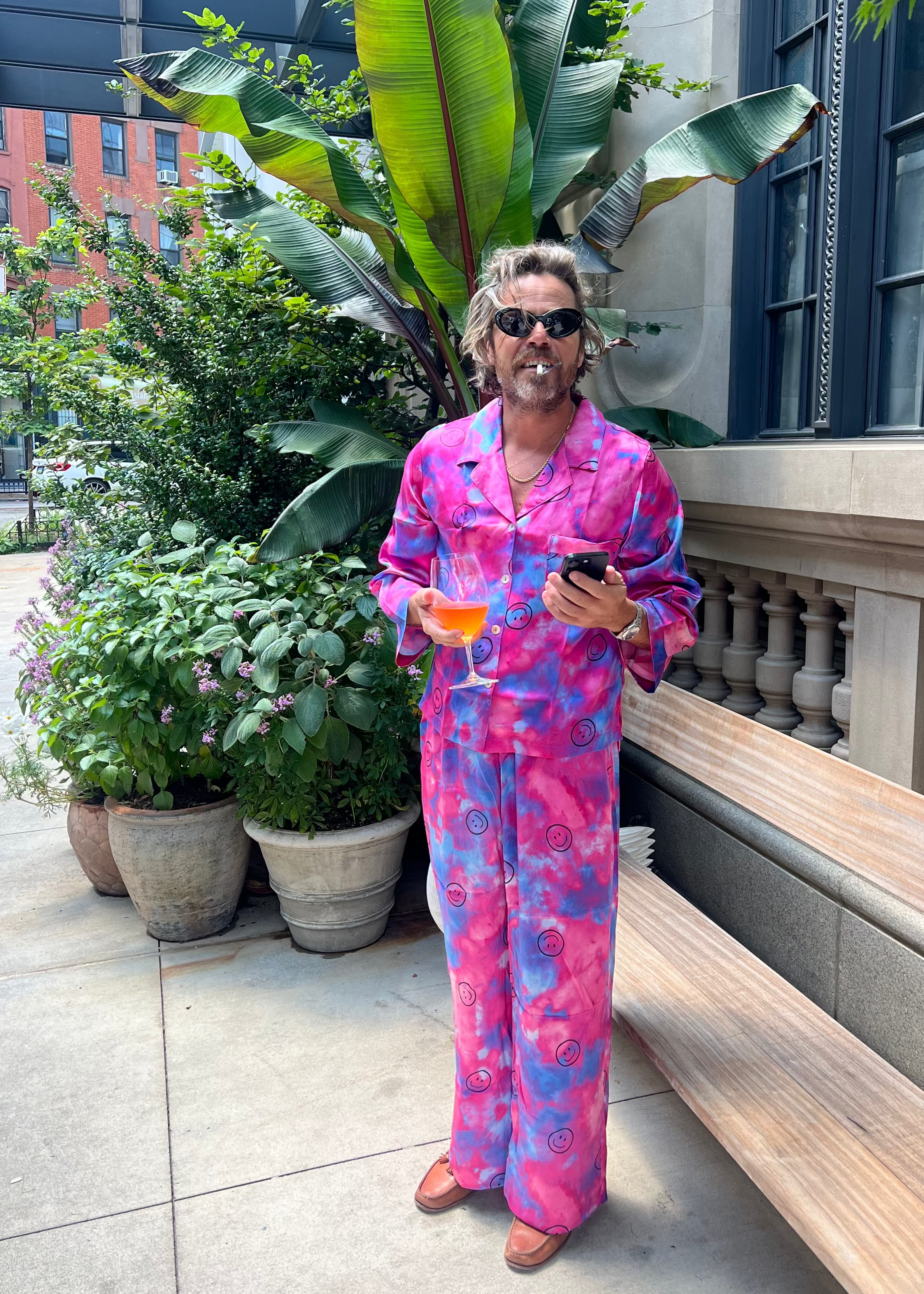 A man wearing a vibrant pink tie dye patterned pajama set stands on an outdoor patio holding a drink and a phone. 